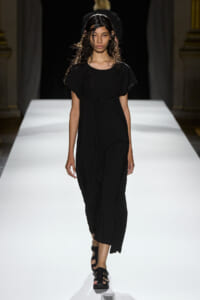Model wearing a long black dress and sandals walks down a white runway in a fashion show.