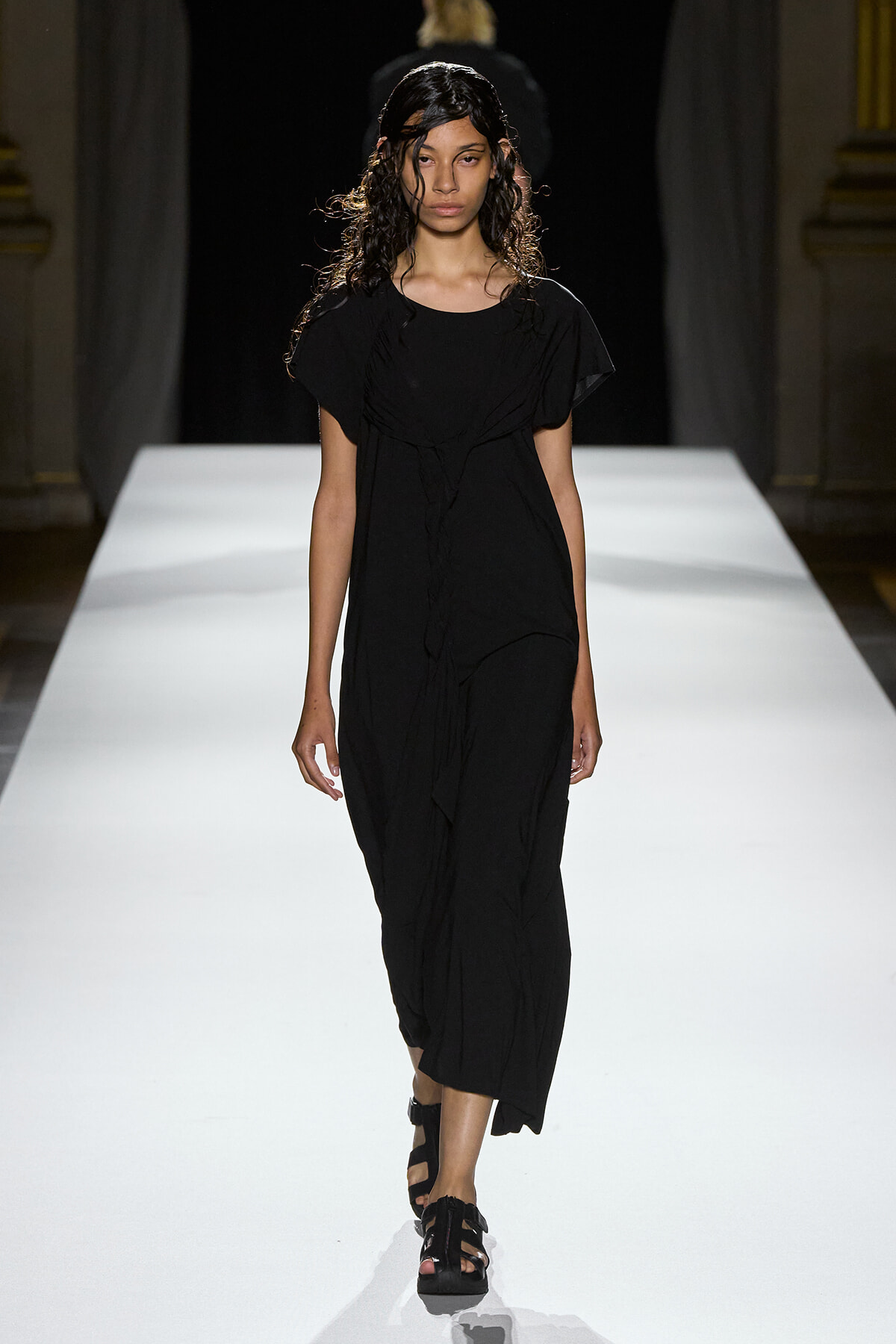 Model wearing a long black dress and sandals walks down a white runway in a fashion show.