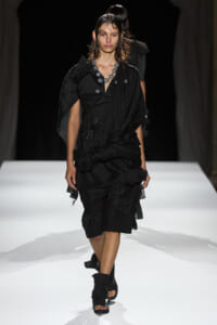 Runway model in a black draped dress with ruching and floral embellishments, paired with a chunky silver necklace and wet-looking hair.