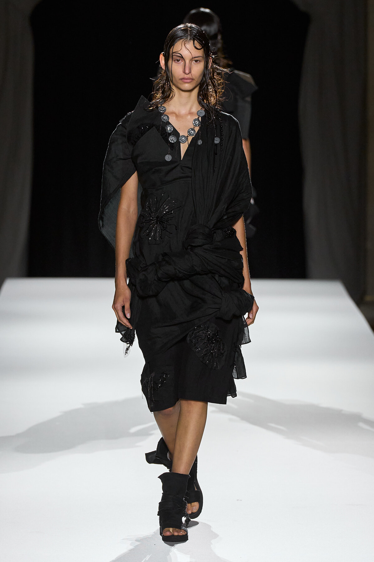 Runway model in a black draped dress with ruching and floral embellishments, paired with a chunky silver necklace and wet-looking hair.