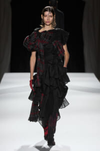 Model walking a runway in a black, multi-layered ruffled gown with a red plaid scarf draped over one shoulder and a red glove on one hand.