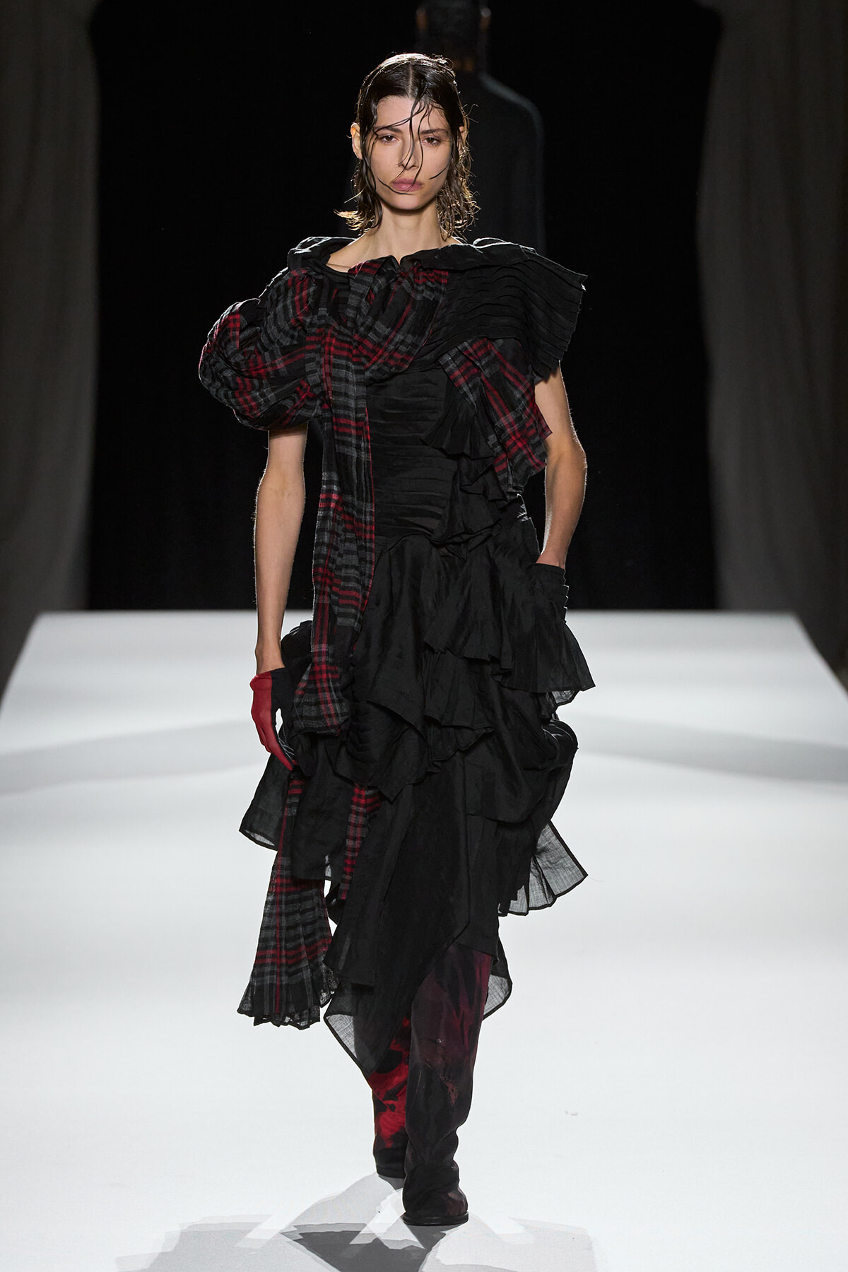 Model walking a runway in a black, multi-layered ruffled gown with a red plaid scarf draped over one shoulder and a red glove on one hand.