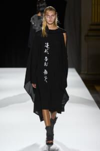 Model walks the runway in a black cape-style dress with vertical white script down the front.