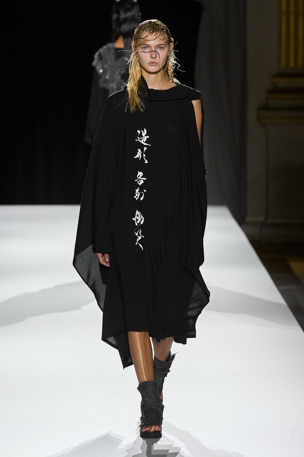 Model walks the runway in a black cape-style dress with vertical white script down the front.