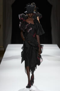 Model walking the runway in a black layered gown with plaid accents and a tall feathered hat.