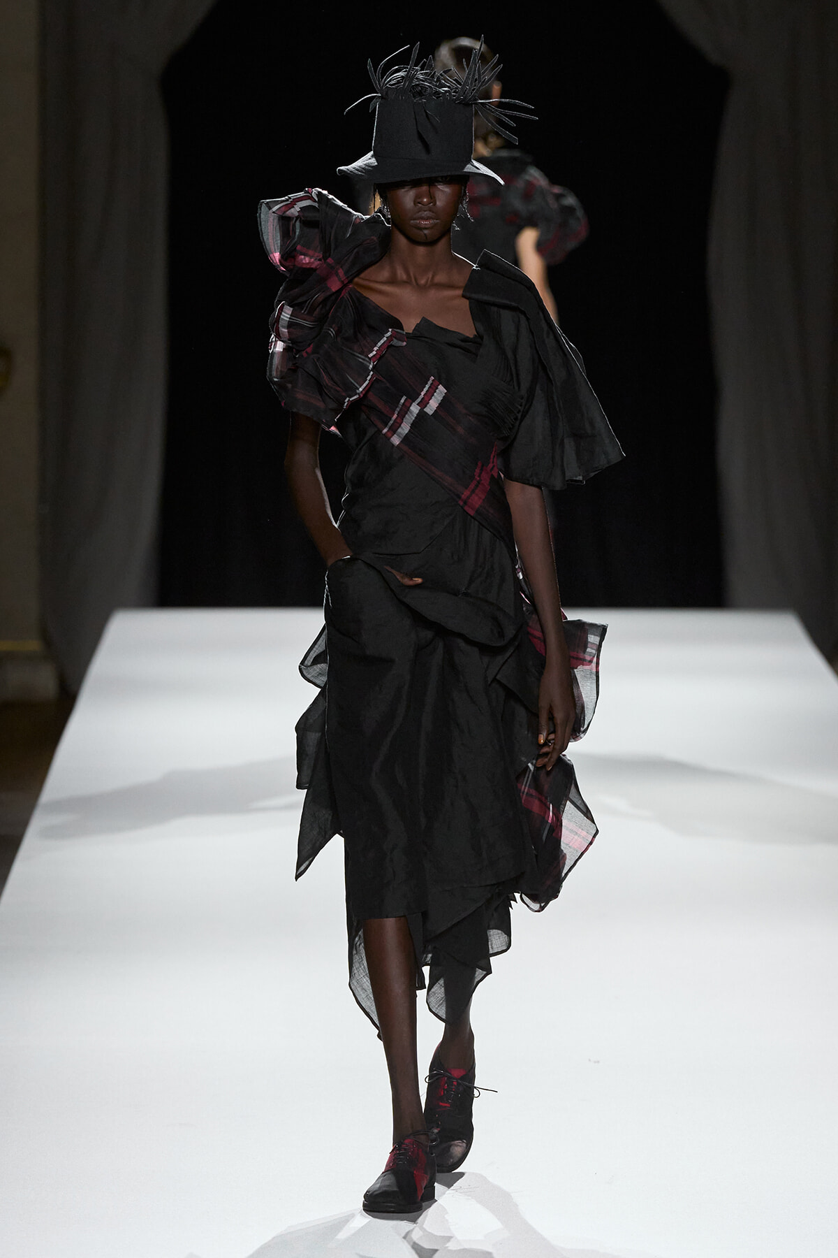 Model walking the runway in a black layered gown with plaid accents and a tall feathered hat.