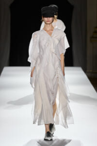 Model walking a white flowing, pleated dress on a runway, wearing a black cap and sneakers.