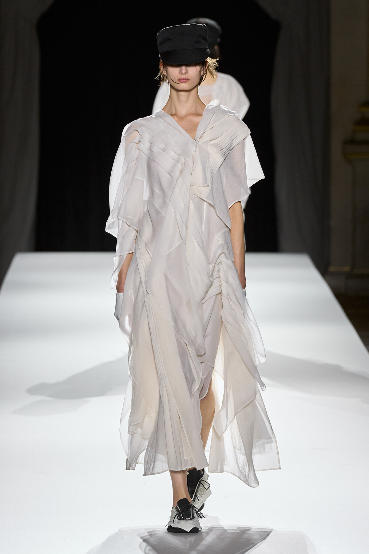 Model walking a white flowing, pleated dress on a runway, wearing a black cap and sneakers.