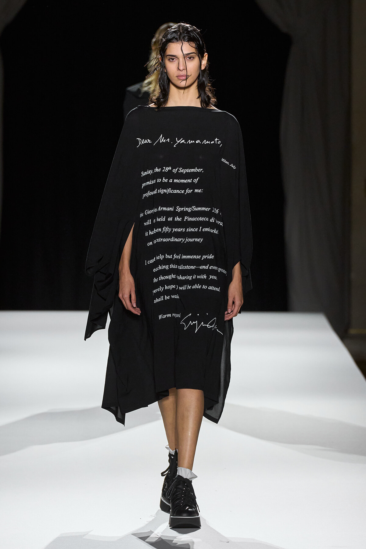 Model walks the runway in a long black dress covered in white handwritten text, pairing with black boots on a white catwalk.