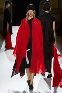 Model on a white runway wearing a bright red draped cape over a black dress and a black cap.