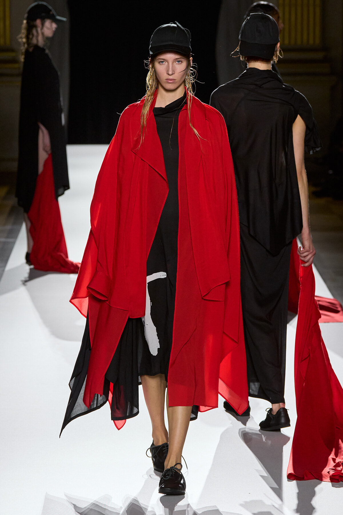 Model on a white runway wearing a bright red draped cape over a black dress and a black cap.