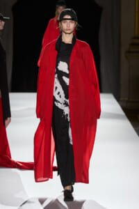 Model on a white runway wearing a red sheer, ankle-length coat over a black-and-white graphic top and black pants, with a backward black cap.
