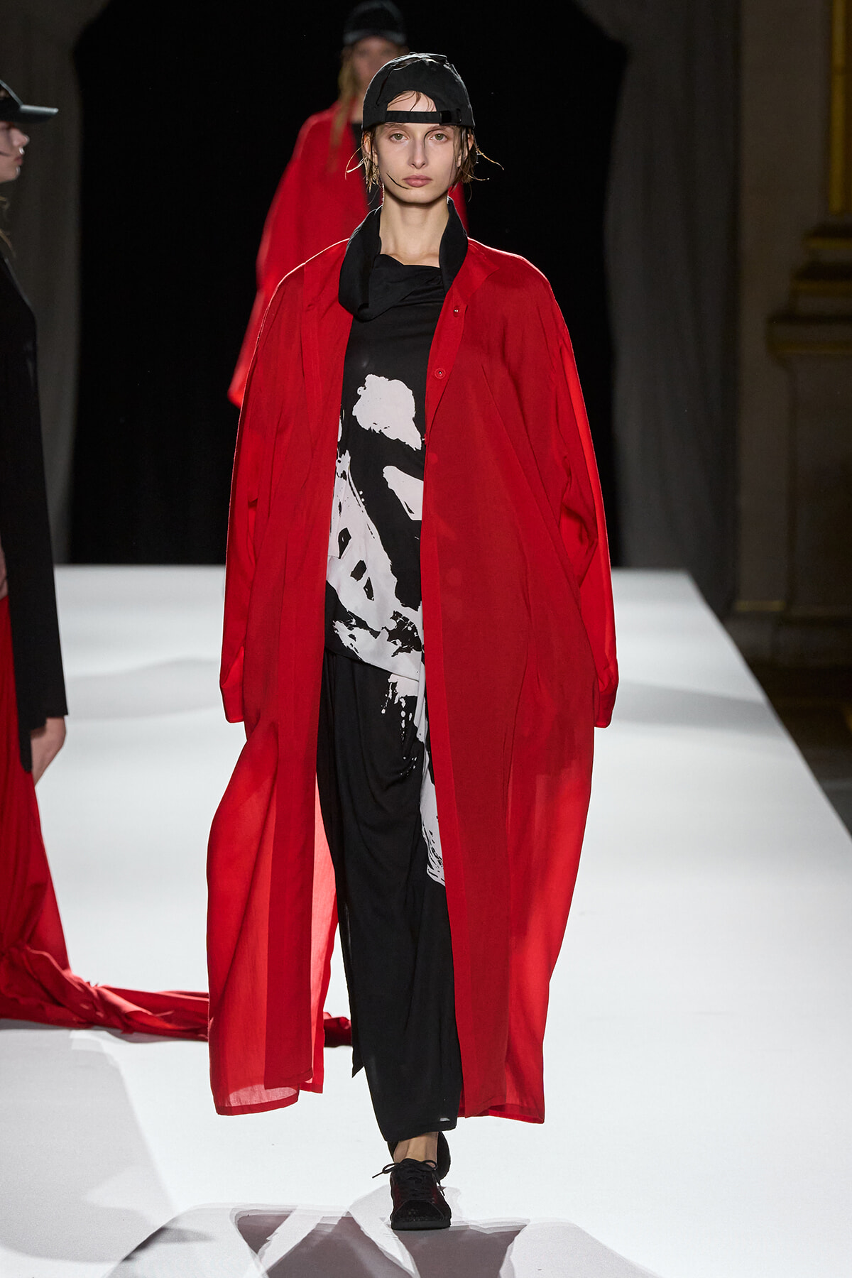 Model on a white runway wearing a red sheer, ankle-length coat over a black-and-white graphic top and black pants, with a backward black cap.