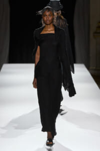 Model walking a fashion runway in a black, off-shoulder dress and matching shawl, on a white catwalk with dark background