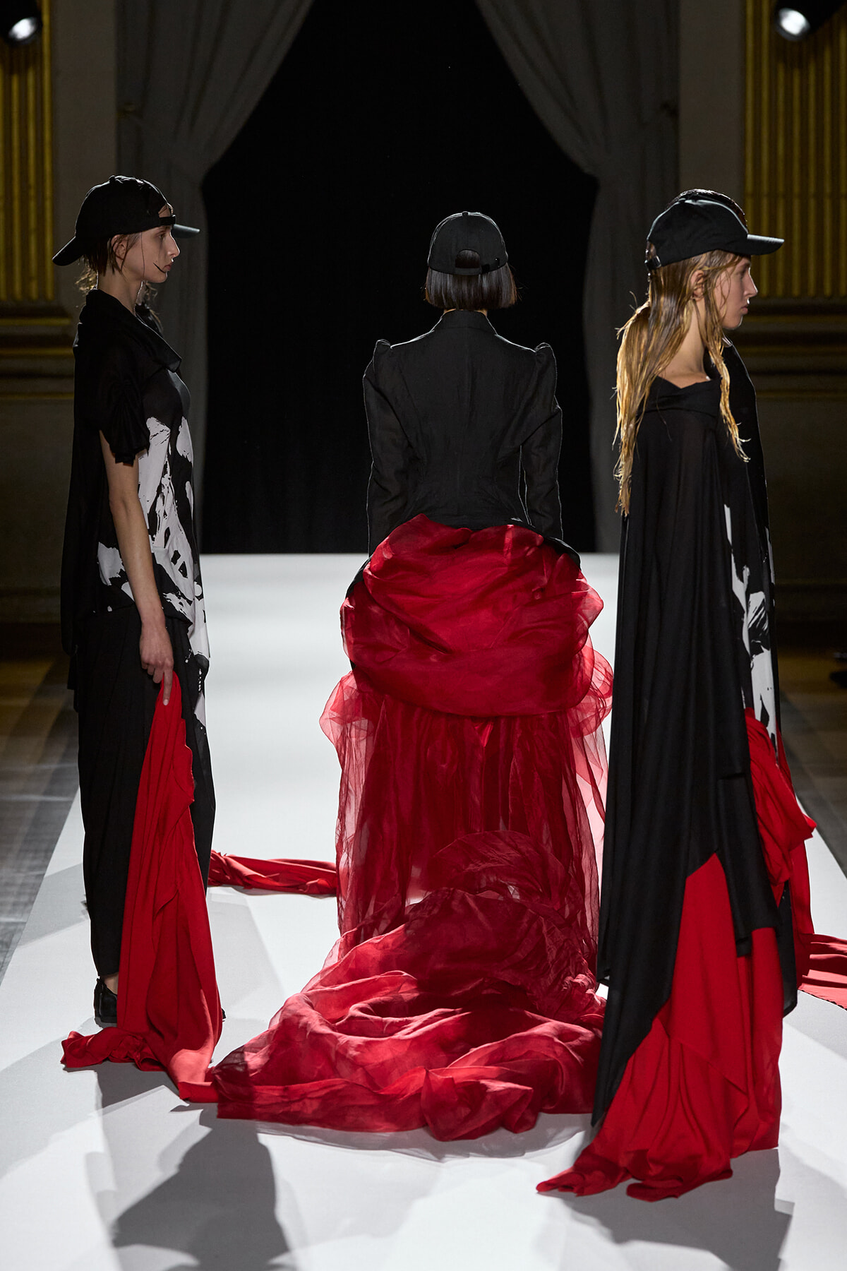 Three models on a fashion runway wearing black outfits with dramatic red train fabrics trailing behind them on a white catwalk.