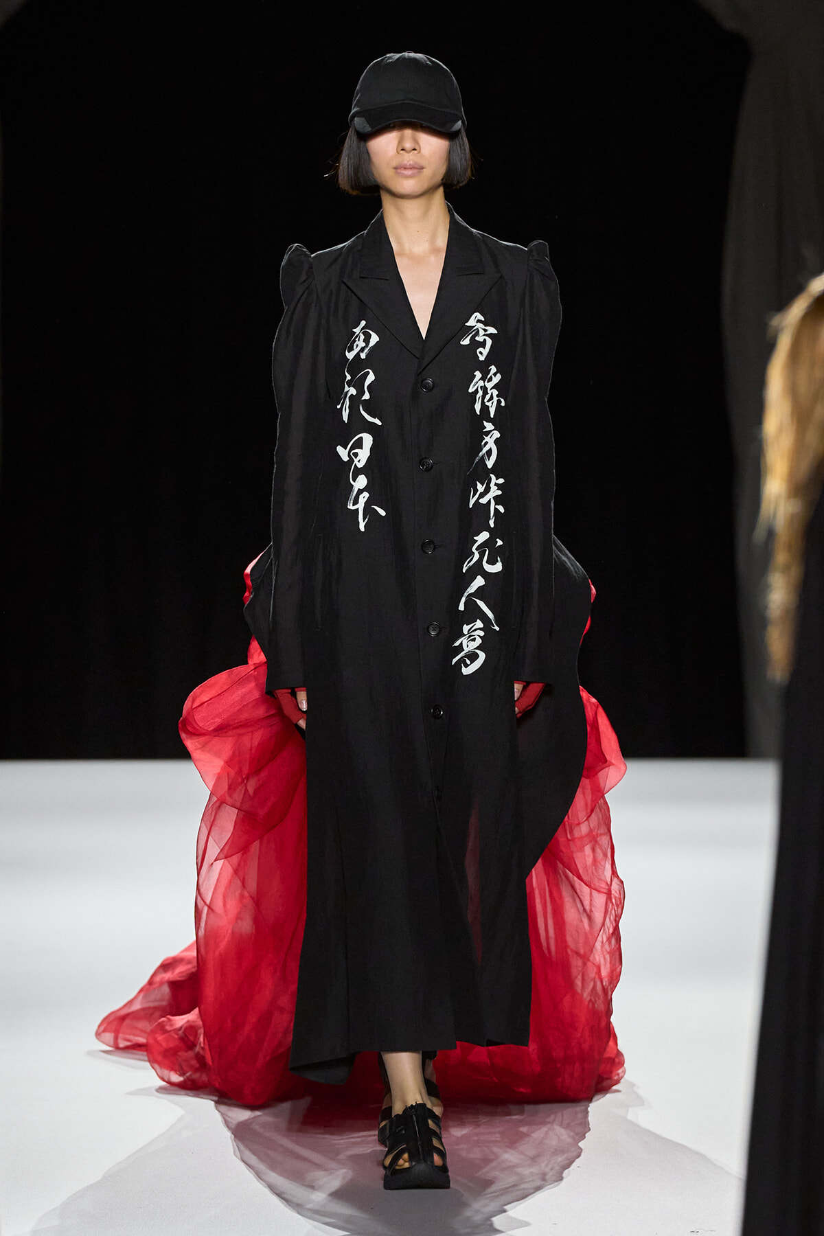 Model walks the runway in a long black coat with white vertical calligraphy, paired with a red sheer cape and a black cap.