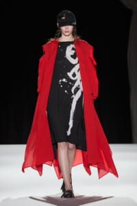Runway model wearing a long red overcoat over a black dress with white abstract print, cap on, walking forward on a white catwalk.