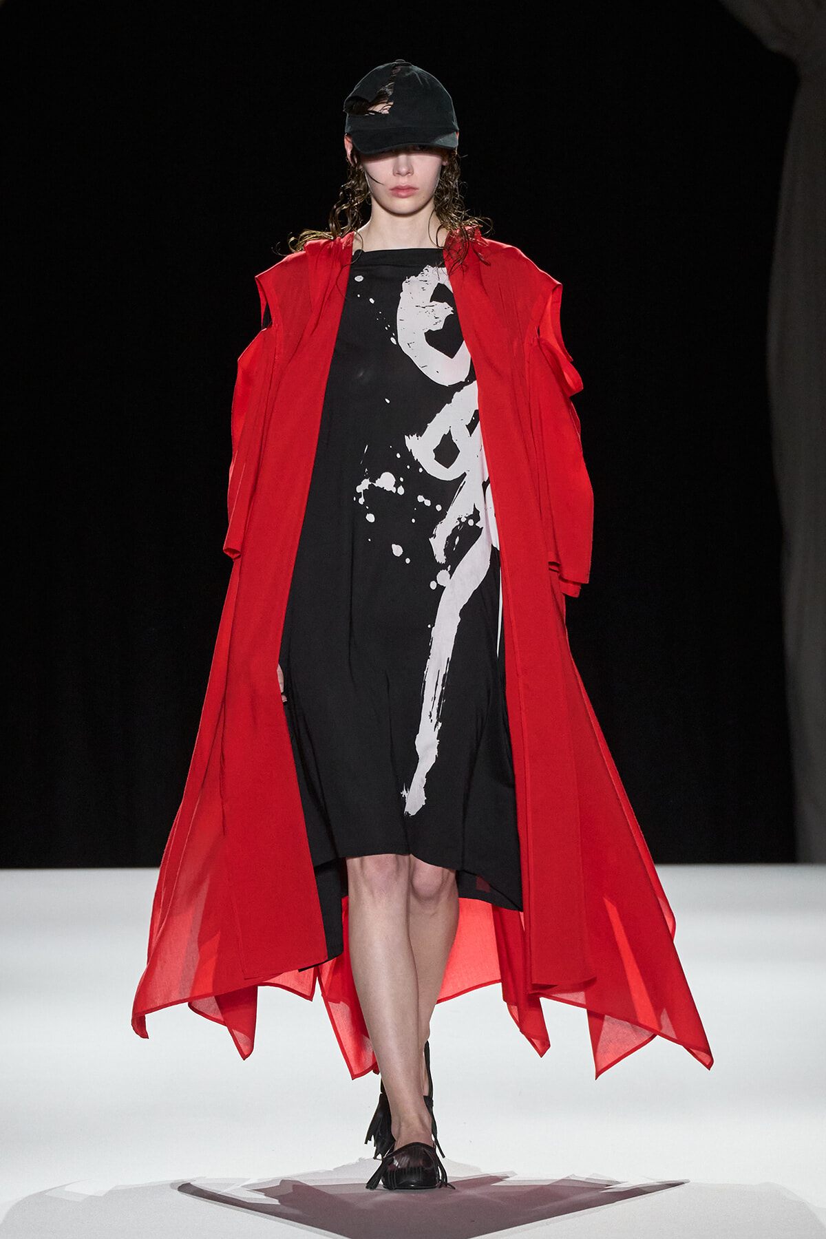 Runway model wearing a long red overcoat over a black dress with white abstract print, cap on, walking forward on a white catwalk.