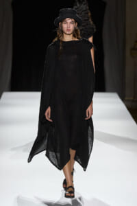 Model walking the runway in a loose black cape dress with a wide brim hat and black sandals.
