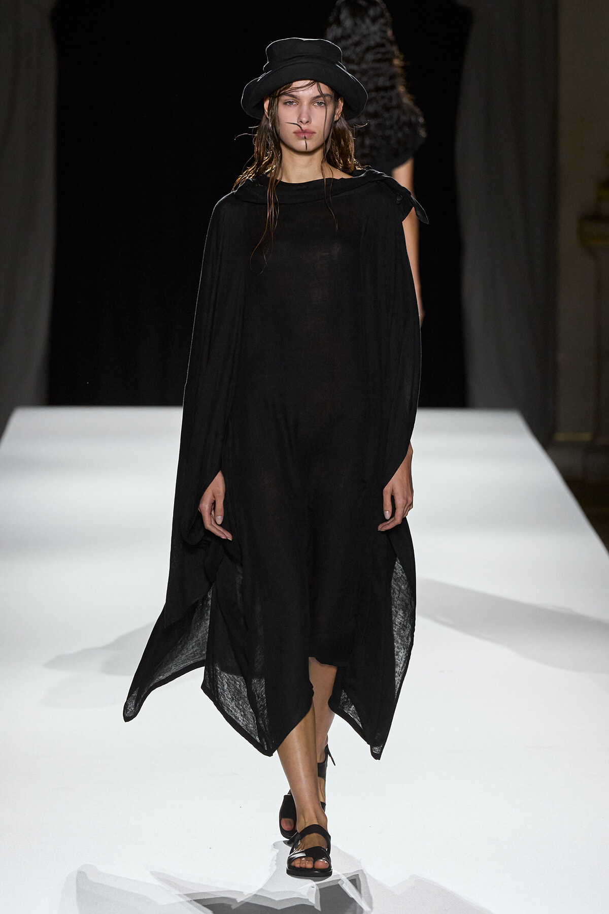 Model walking the runway in a loose black cape dress with a wide brim hat and black sandals.