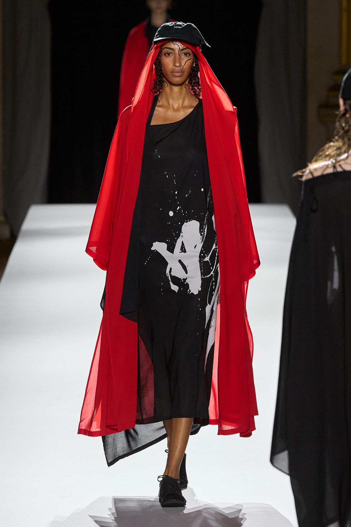 Model walks the runway in a black asymmetrical dress with a white abstract splash print, overlaid by a flowing red sheer cape and a black cap.