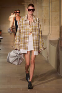 Model on a runway wearing an oversized beige plaid jacket over a white dress, with dark sunglasses and a large gray gathered handbag.