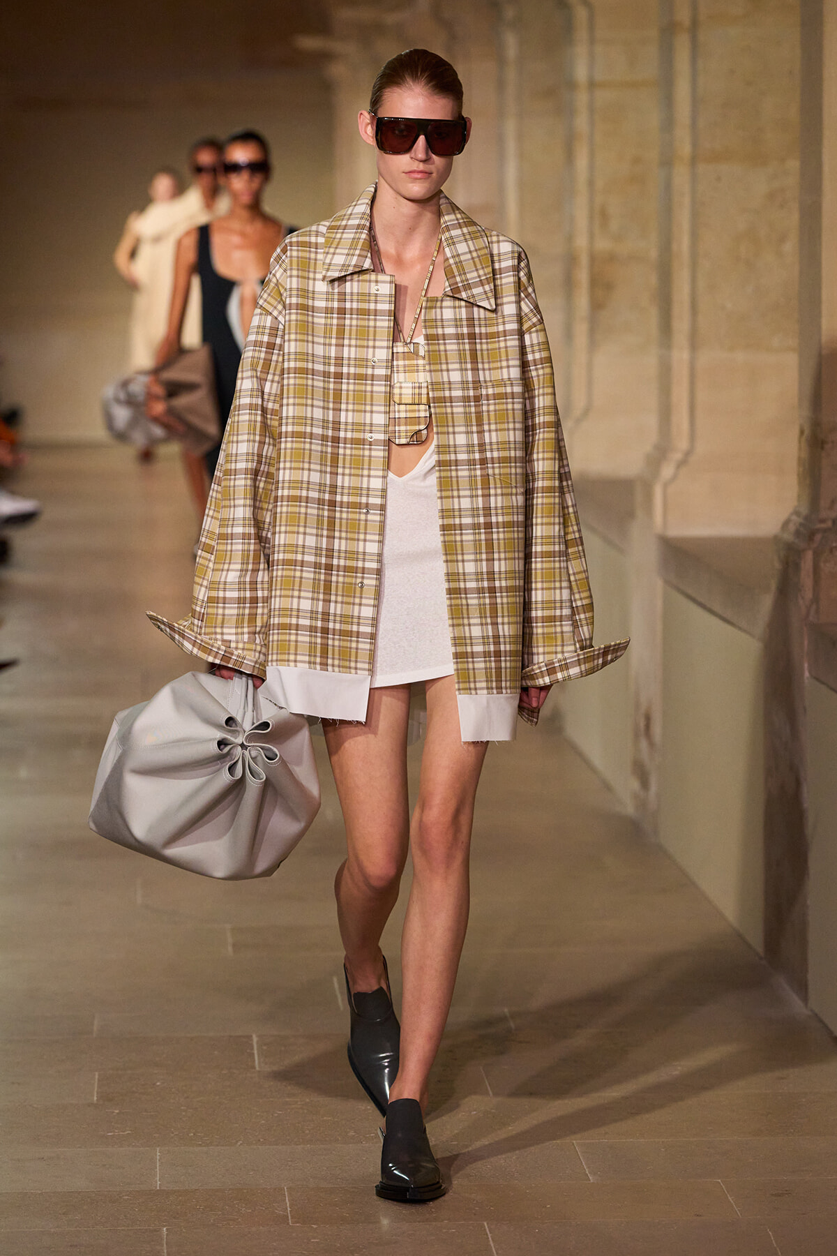 Model on a runway wearing an oversized beige plaid jacket over a white dress, with dark sunglasses and a large gray gathered handbag.