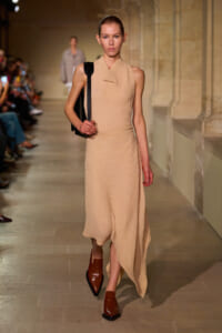 Model walks the runway in a sleeveless beige knit dress with an asymmetrical, draped hem, carrying a black shoulder bag and wearing brown shoes.