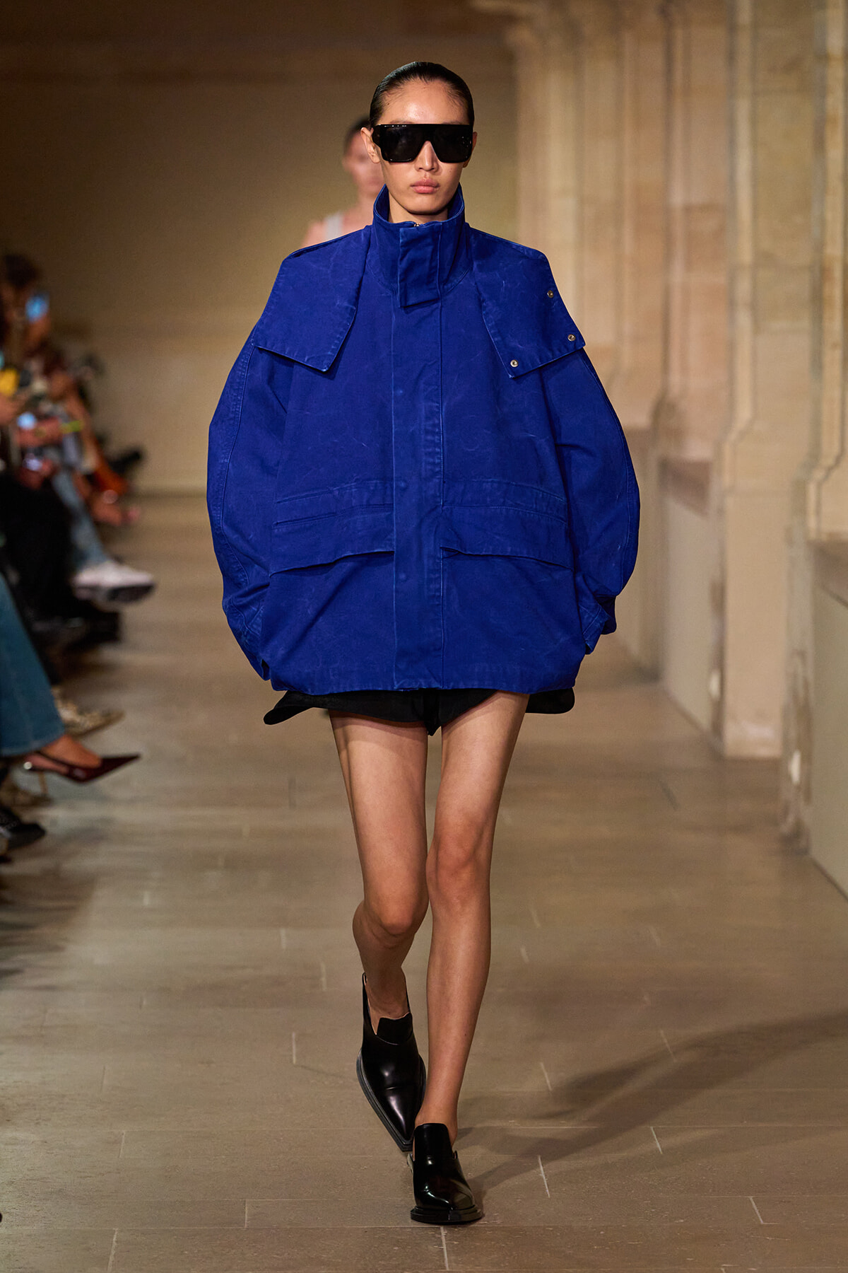 Model on a runway wearing an oversized royal blue jacket, black shorts, and black loafers with oversized sunglasses.