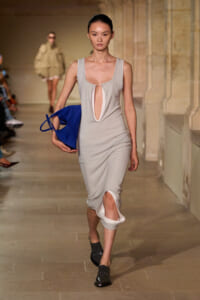 Model walking the runway in a pale gray sleeveless dress with a large front keyhole cutout, carrying a royal blue handbag and wearing black shoes.