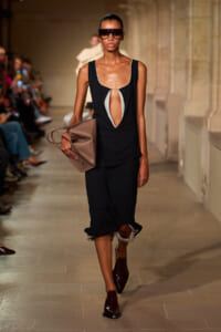 Model walks a runway in a black sleeveless dress with a front cutout, oversized dark sunglasses, burgundy shoes, and a taupe handbag.