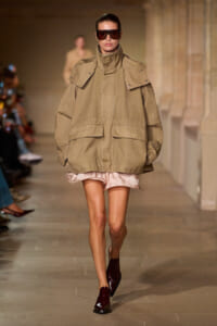 Model walks a fashion runway wearing an oversized khaki parka with a high collar and large flap pockets, paired with burgundy boots and dark sunglasses.