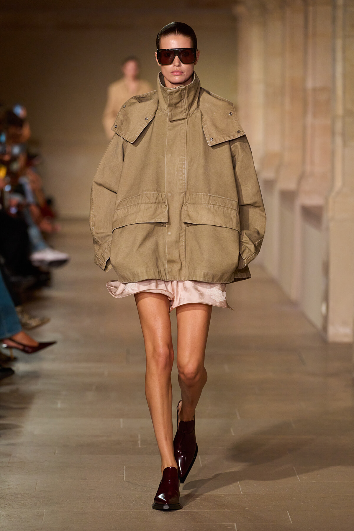 Model walks a fashion runway wearing an oversized khaki parka with a high collar and large flap pockets, paired with burgundy boots and dark sunglasses.