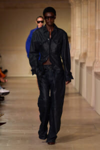 Model walks a runway in a black leather jacket and matching wide-leg pants, wearing dark sunglasses. A second model in blue appears blurred in the background.