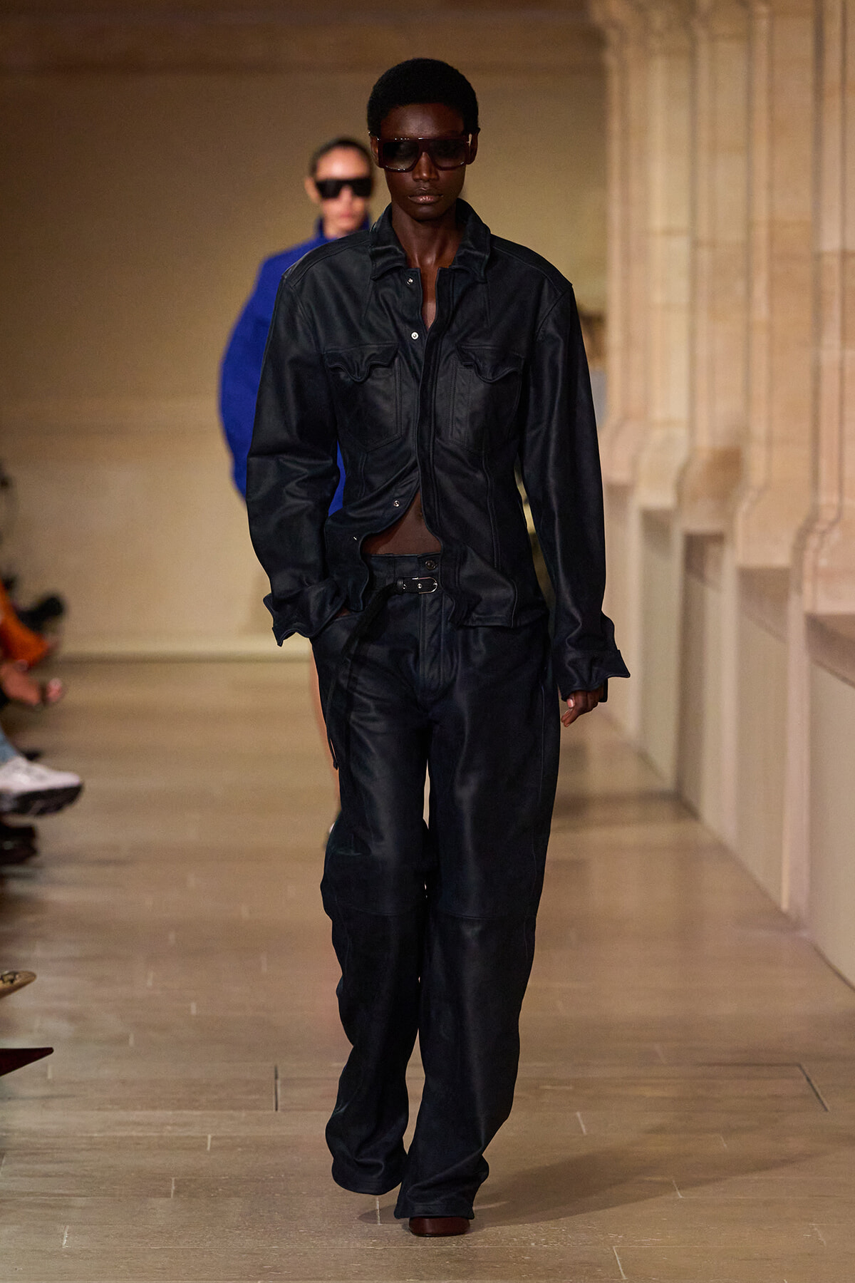 Model walks a runway in a black leather jacket and matching wide-leg pants, wearing dark sunglasses. A second model in blue appears blurred in the background.