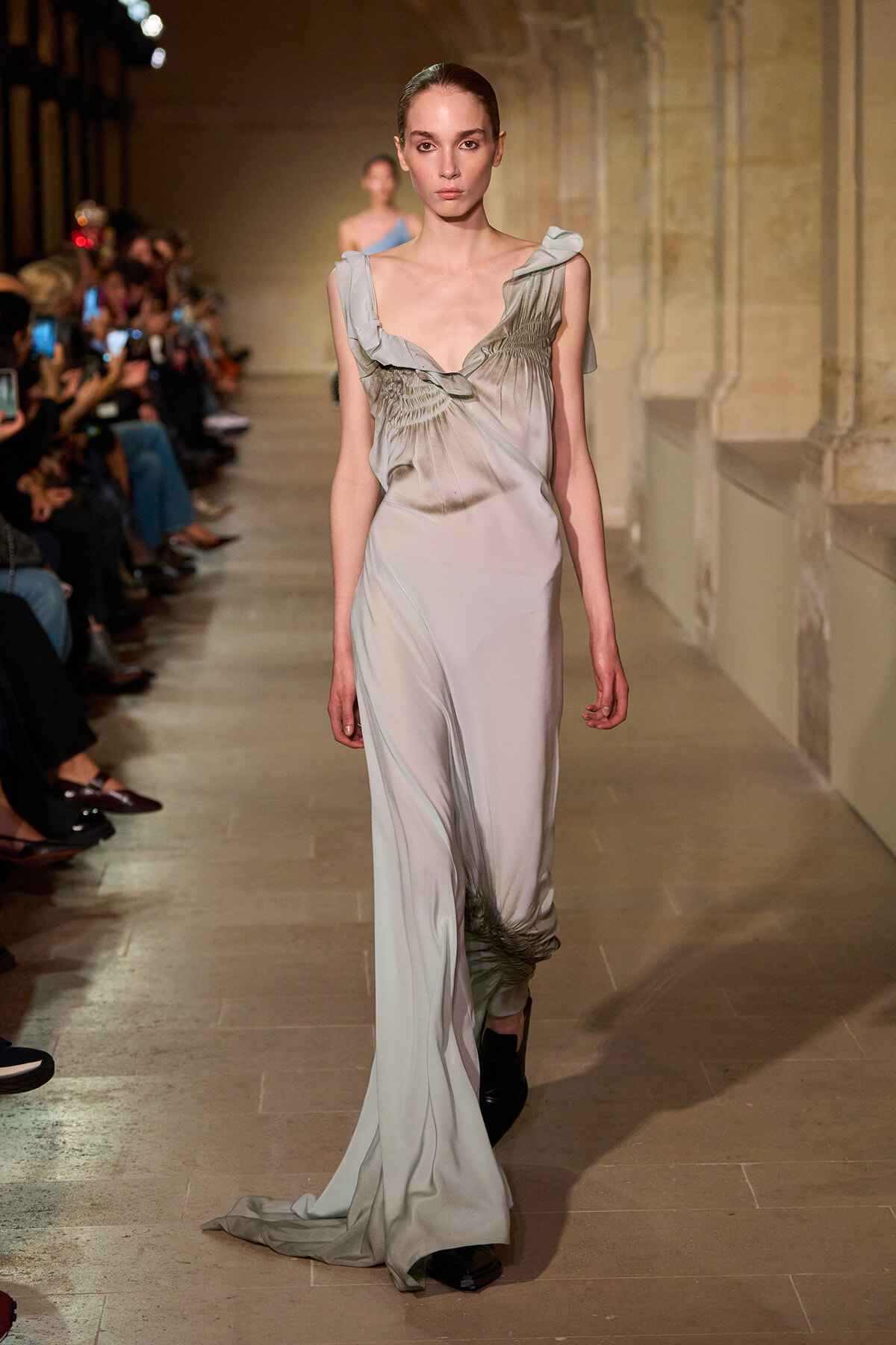 Model walks a pale gray gown with gathered bodice down a fashion runway, audience seated along the sides.