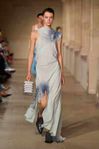 Model walking a runway in a pale blue-gray draped gown with a gathered detail at the chest, holding a metallic pleated clutch.