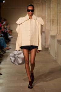 Model on a runway wearing an oversized beige cape coat with a large collar, black shorts, burgundy ankle boots, oversized sunglasses, and a gray handbag with a sculptural flower-like knot.
