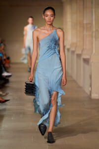 Model in a light blue one-shoulder dress with a ruffled side walking a runway, holding a black stackable clutch.