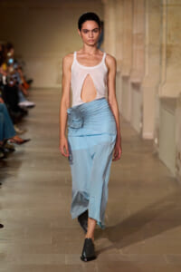 Model walking runway in a white sleeveless top with a front cutout and a light blue ruched skirt with a flower detail at the hip.