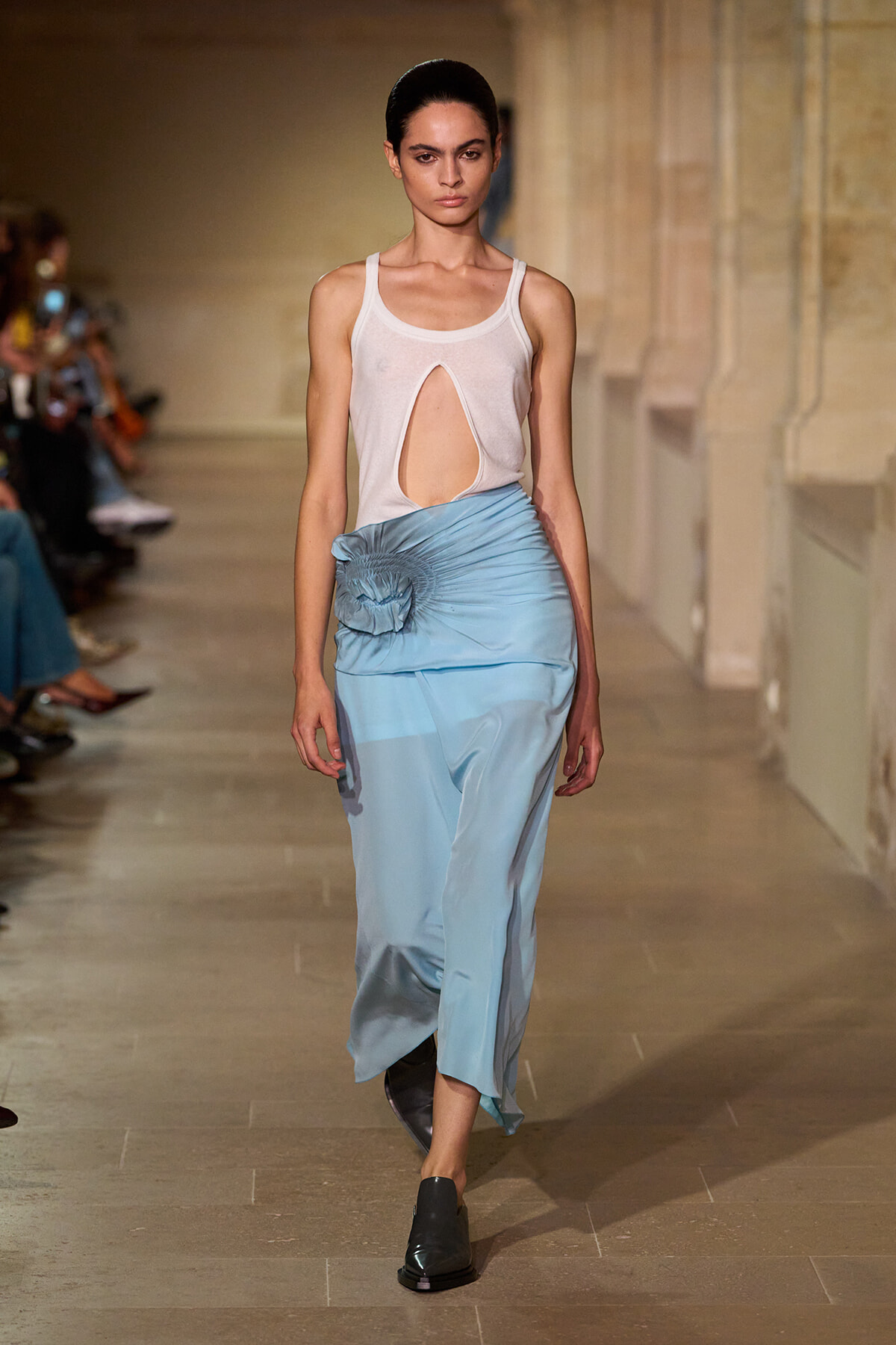 Model walking runway in a white sleeveless top with a front cutout and a light blue ruched skirt with a flower detail at the hip.
