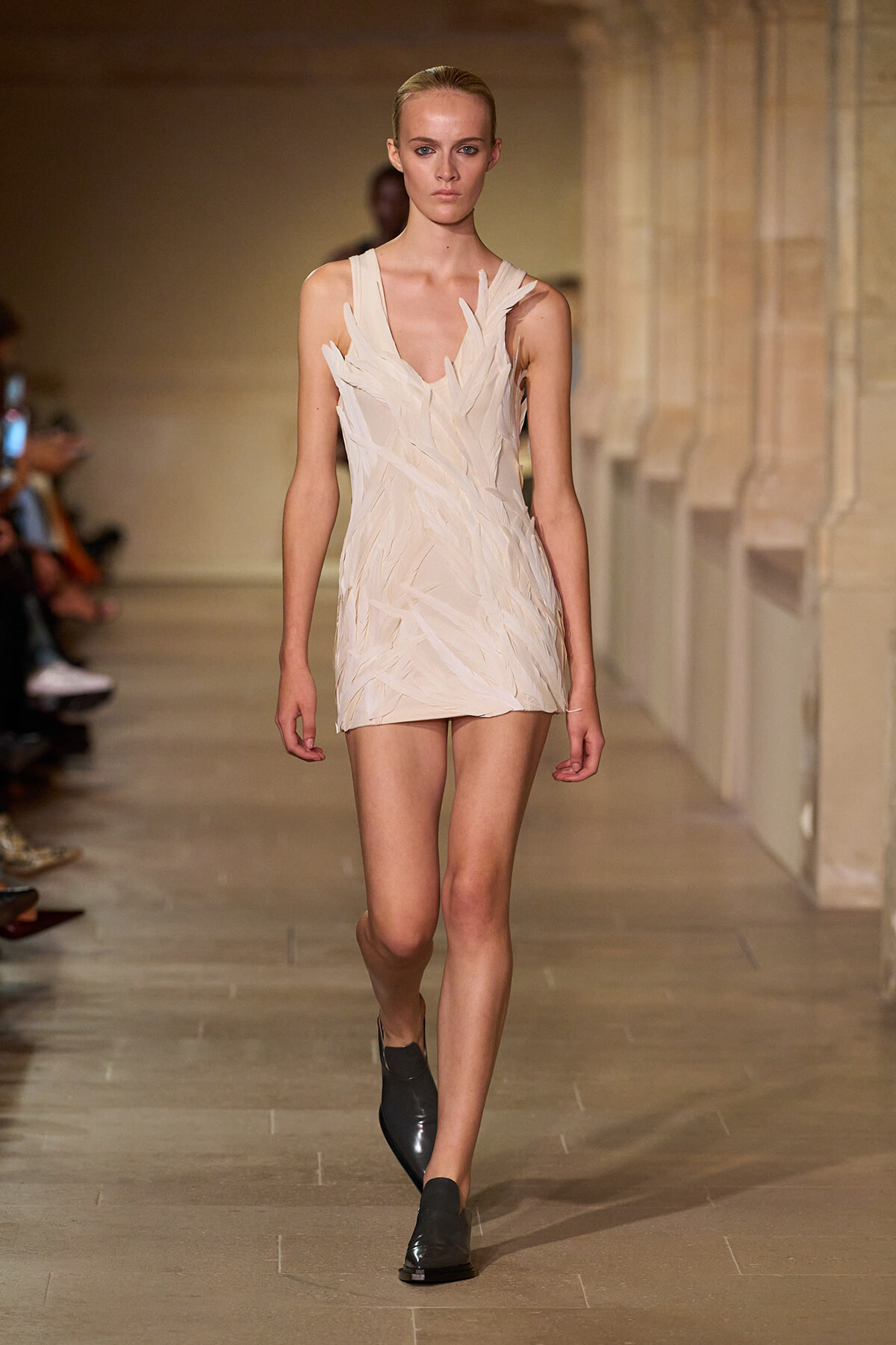 Model walking the runway in a cream leaf-textured mini dress with black shoes on a fashion catwalk.