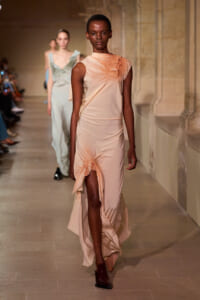 Model walks the runway in a peach satin one-shoulder gown with ruched bodice and asymmetrical drape near the shoulder and hip.
