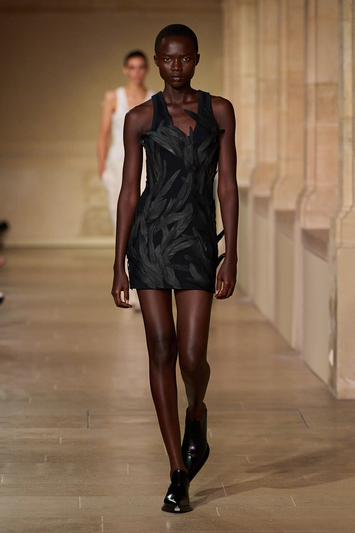 Model on a runway wearing a black feather-embellished mini dress with black ankle boots.