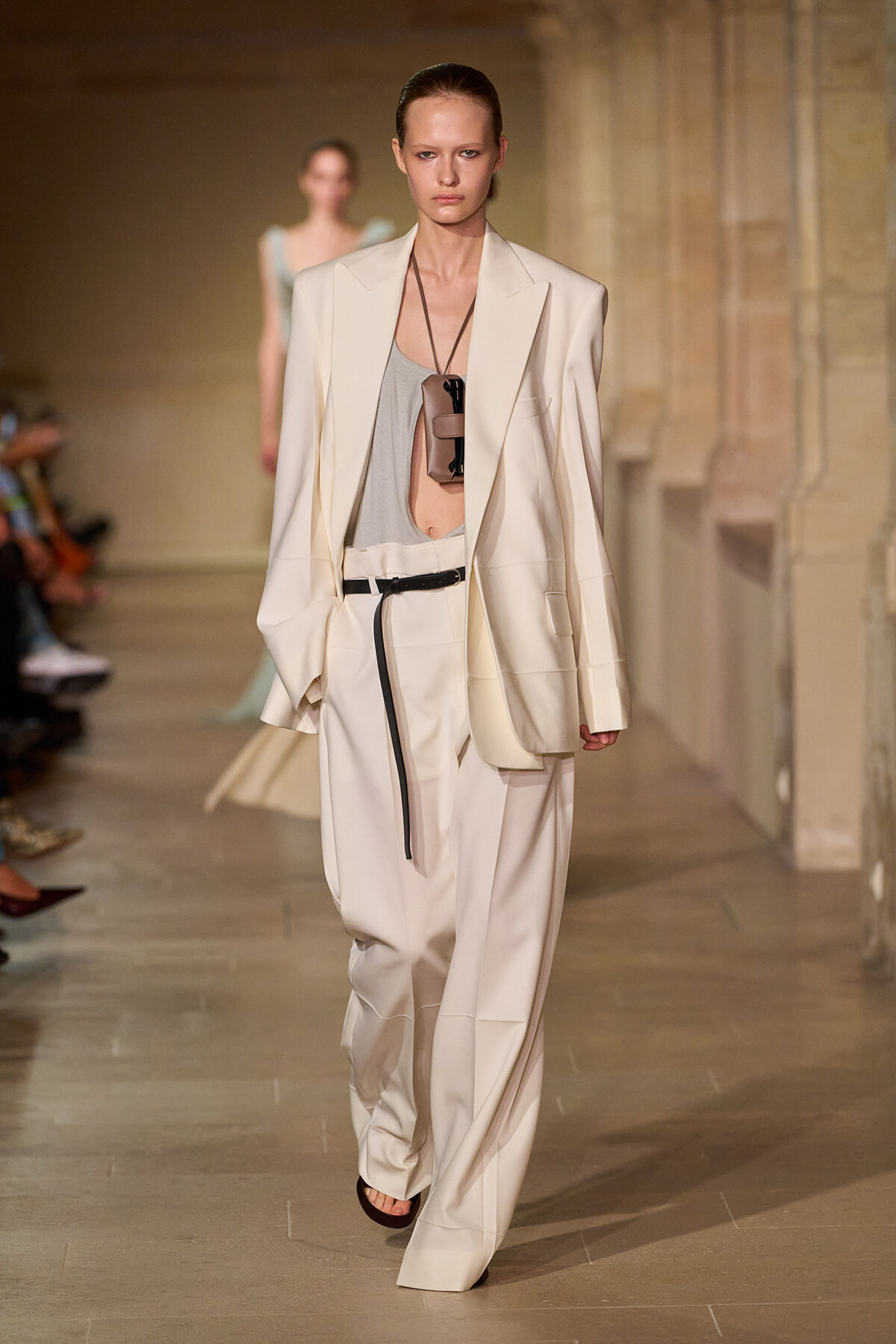 Model on runway wearing an oversized cream suit with a loose blazer, high-waisted wide-leg trousers, and a gray top; black belt and accessory hanging at chest.