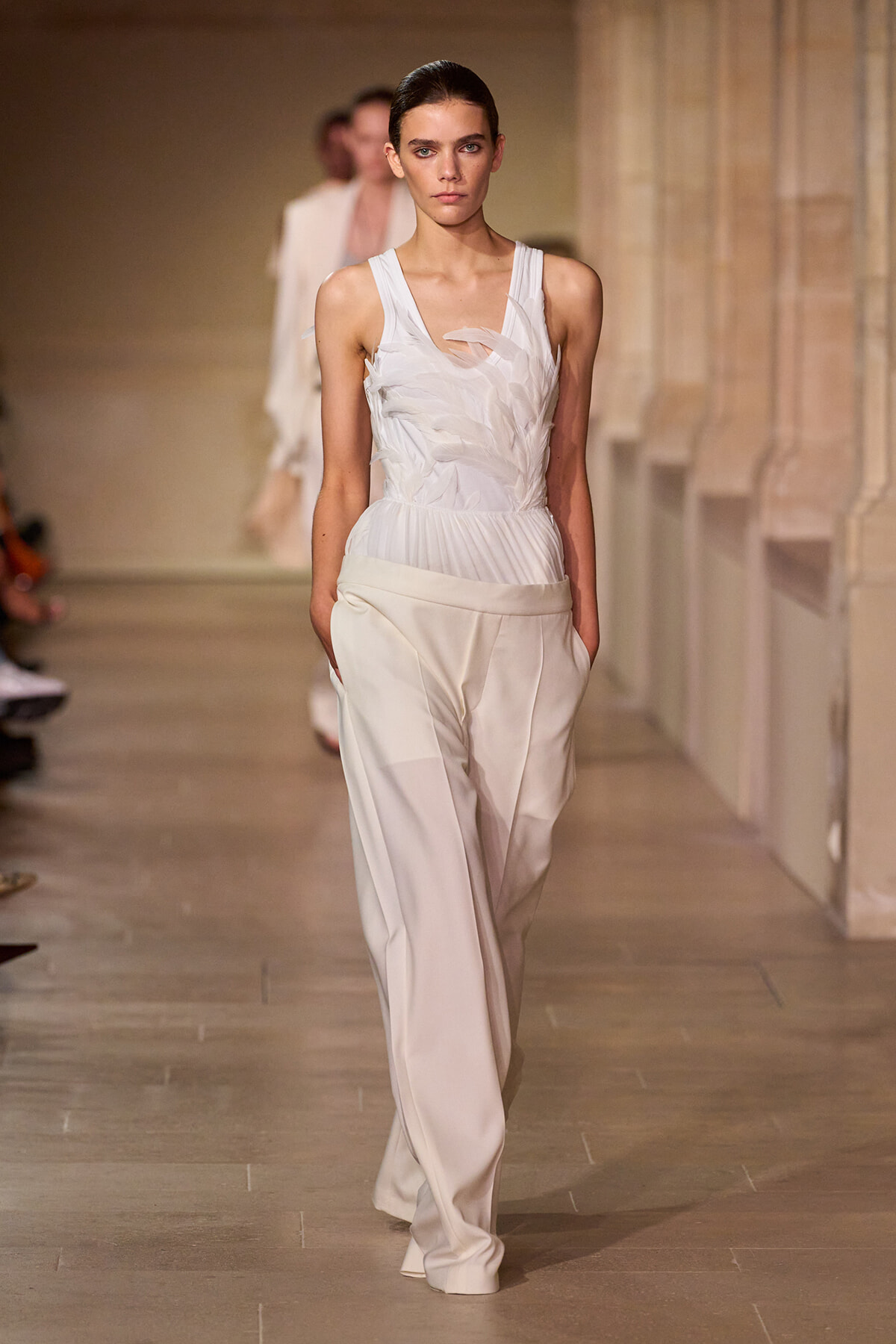 Model walks a runway in an all-white ensemble: textured sleeveless top and loose ivory pants with pockets.