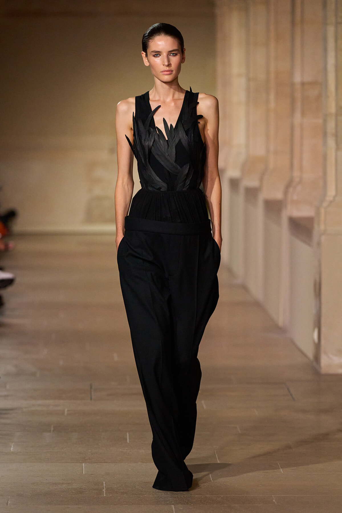 Model walking a runway in a sleeveless black dress with feather-like cutouts at the bodice, high waist, and wide-legged pants.