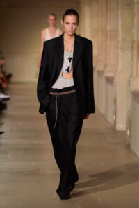 Model walking a runway in a black oversized blazer over a light gray top, black wide-leg pants, and a brown belt; bold white pendant necklace hanging at chest level.
