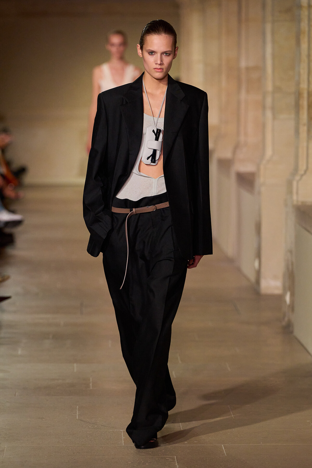 Model walking a runway in a black oversized blazer over a light gray top, black wide-leg pants, and a brown belt; bold white pendant necklace hanging at chest level.