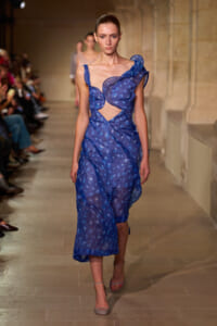 Model walking a runway in a blue patterned cutout evening dress and beige heels, audience on sides.